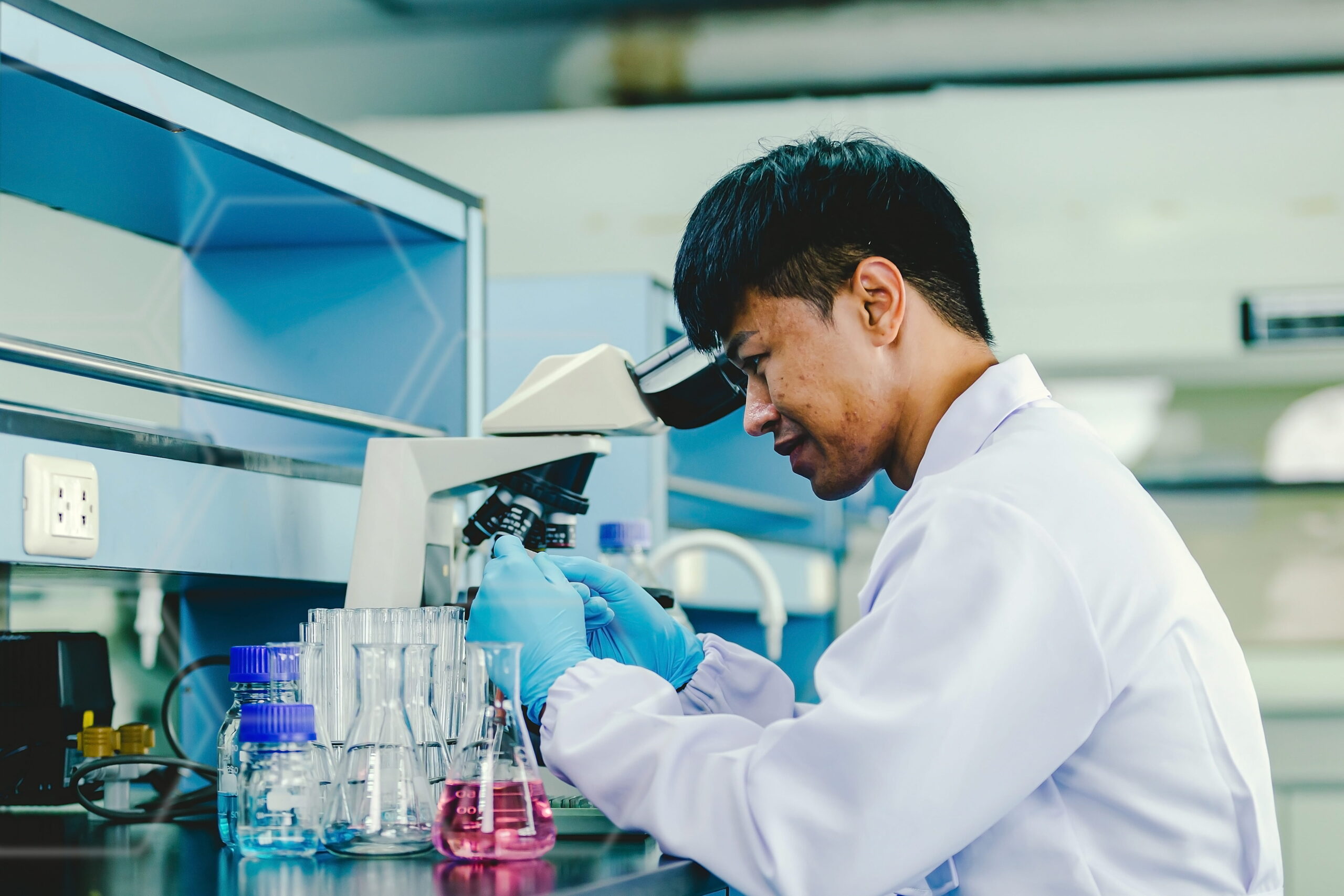 Asian man lab technician protective glasses gloves sits microscope laboratory 1 scaled